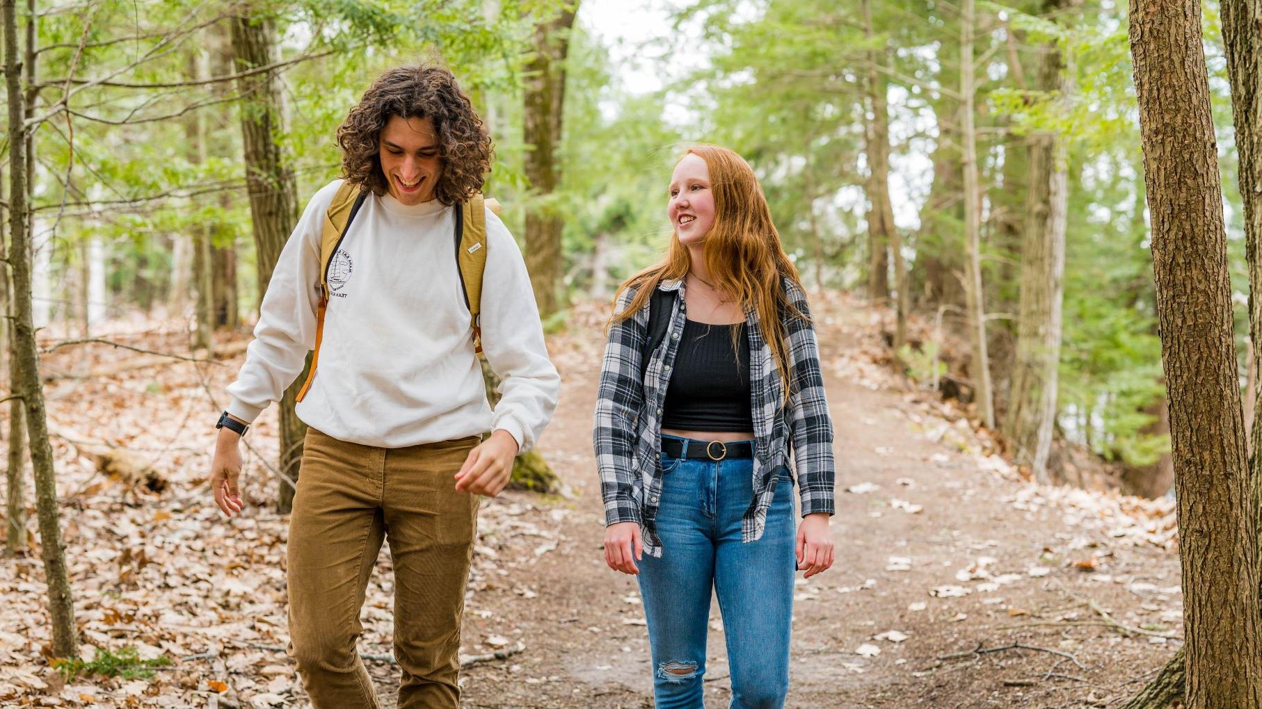 Two students smiling and in conversation as they walk toward the camera in a wooded trail.